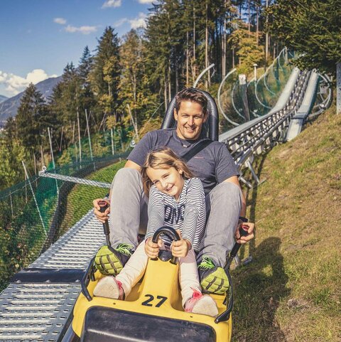 Ein Mann und ein Mädchen fahren mit einer Sommerrodelbahn.