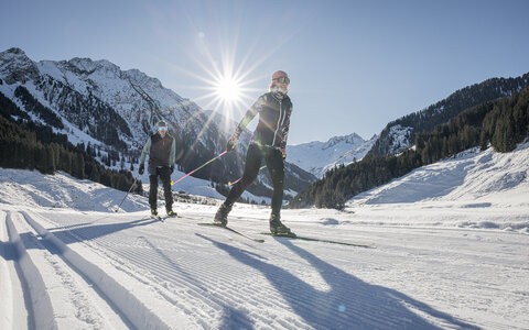 Zwei Langläufer:innen ziehen bei Sonnenschein durch eine verschneite Berglandschaft mit gespurter Loipe.