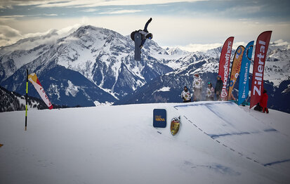 Snowboarder springt über eine Schanze mit Bergkulisse im Hintergrund.