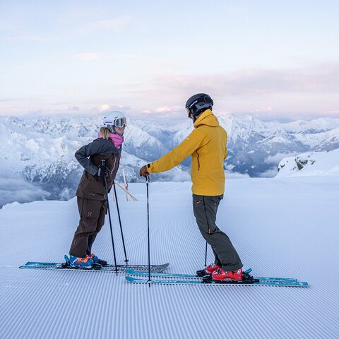 Zwei Skifahrer stehen auf der Piste und unterhalten sich vor einer verschneiten Berglandschaft.