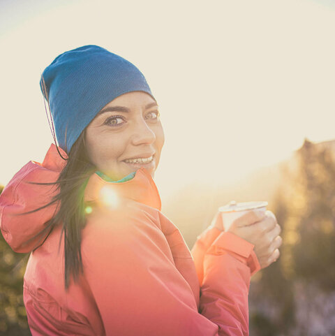 Eine Frau steht, mit einer Tasse in der Hand, am Gipfelkreuz
