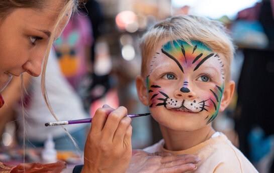 Foto wo eine Frau einen kleinen Jungen einen bunten Tiger ins Gesicht schminkt