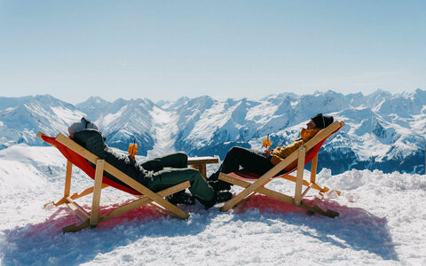 Zwei Personen entspannen auf Liegestühlen im Schnee vor einer verschneiten Berglandschaft.