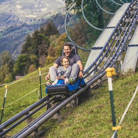 Ein Mann und ein Mädchen fahren mit einer Sommerrodelbahn.