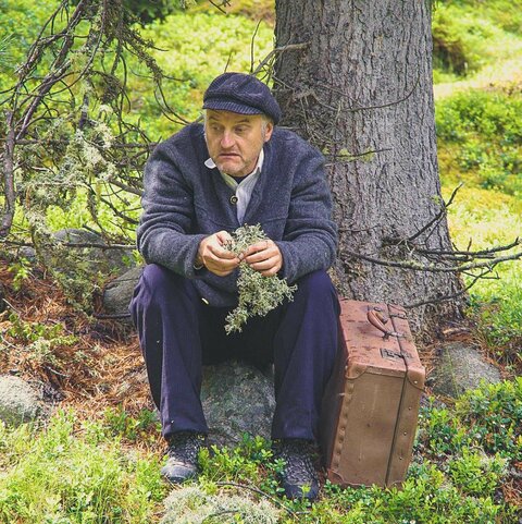 Ein dunkelblau gekleideter Mann sitzt im Wald, mit einem großen braunen Koffer neben sich