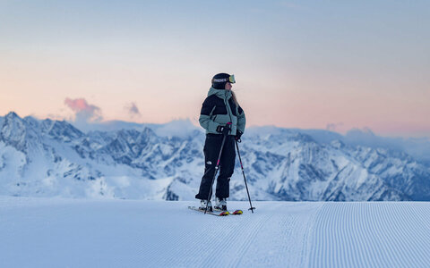 Skifahrer steht auf einer präparierten Skipiste vor einer verschneiten Berglandschaft.