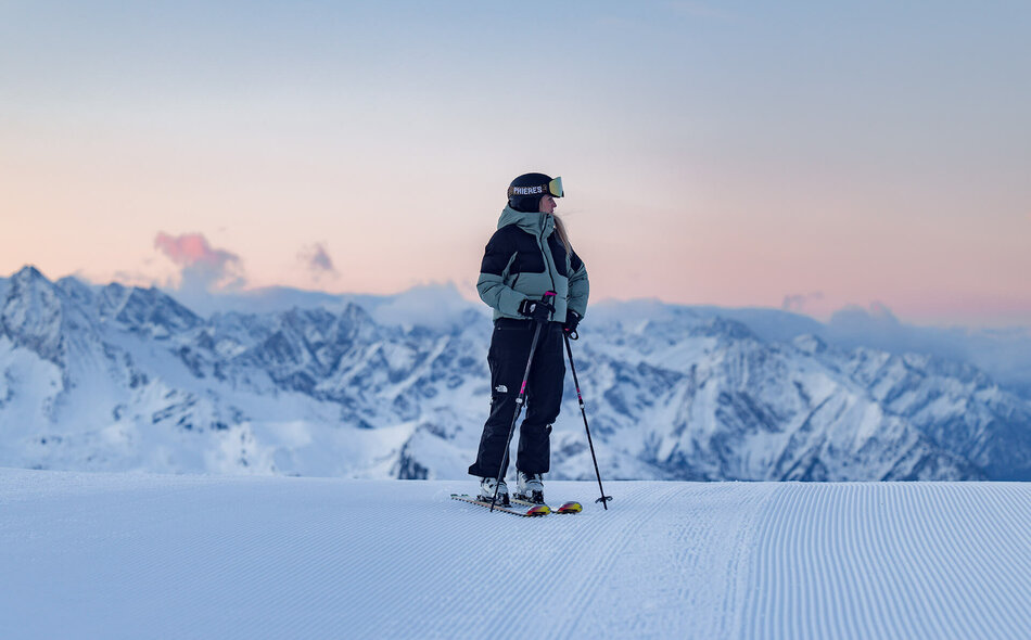 Skifahrer steht auf einer präparierten Skipiste vor einer verschneiten Berglandschaft.