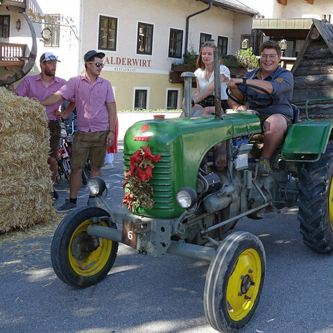 2 Leute sitzen auf Traktor mit angehaengter Holzhuette, daneben Zuschauer und Deko