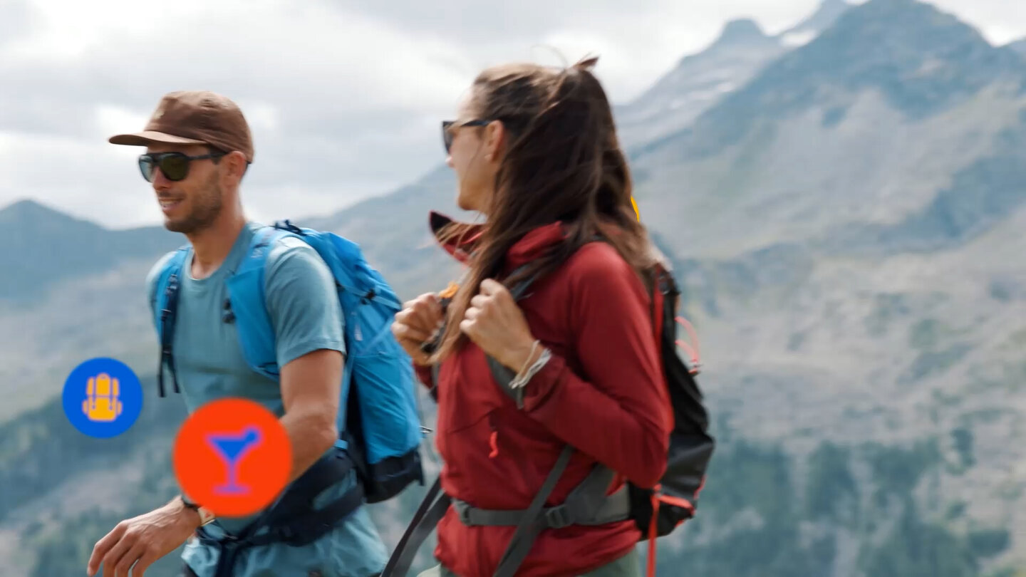 Ein Mann und eine Frau wandern mit Rucksäcken durch eine sonnige Berglandschaft.