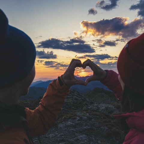 Ein Mann und eine Frau formen ein Herz mit ihren Händen und beobachten einen Sonnenuntergang
