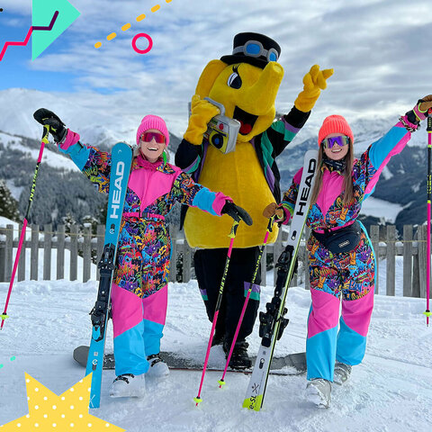 Zwei in Pink gekleidete Skifahrerinnen stehen lächelnd neben dem Maskottchen Funty