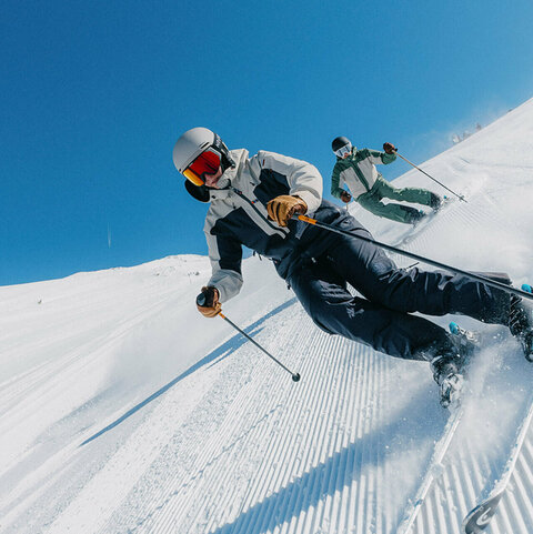 Zwei Skifahrer ziehen Schwünge auf einer fisch präparierten Skipiste unter blauem Himmel.