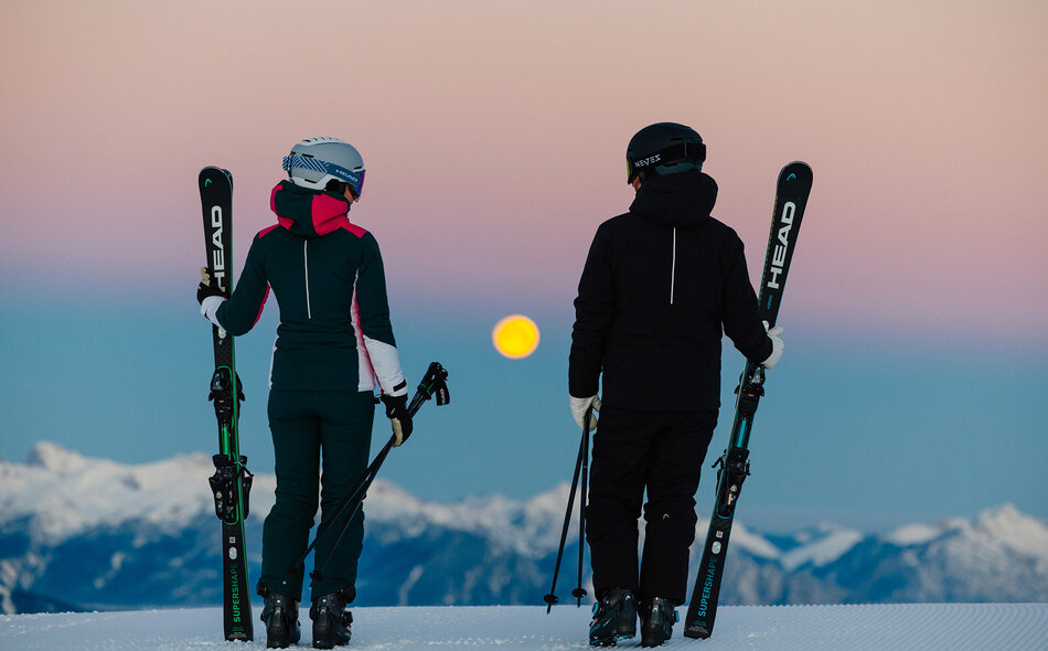 Zwei Skifahrer:innen stehen auf einer verschneiten Bergkuppe und schauen bei Dämmerung auf den aufgehenden Vollmond.