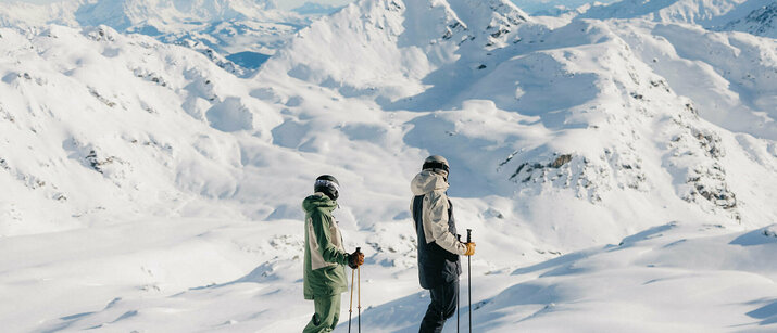 Zwei Skifahrer stehen auf einer präparierten Skipiste vor einer verschneiten Berglandschaft.