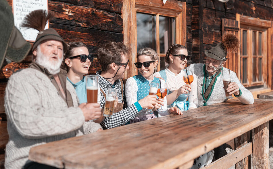 Drei Männer und drei Frauen in Trachtenkleidung trinken Bier und Aperol bei einer Hütte