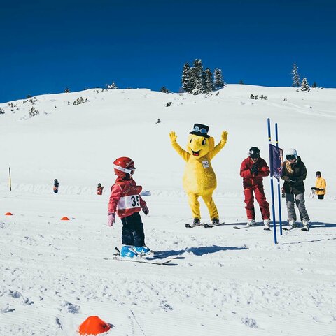 Kleines Kind auf Skiern wird von dem Maskottchen Funty und zwei Erwachsenen angefeuert