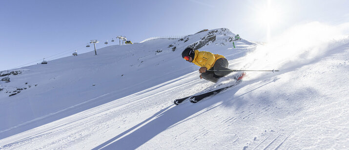 Ein Skifahrer in gelber Jacke fährt schnell eine sonnige, verschneite Piste hinab, mit Bergbahn und Bergen im Hintergrund.