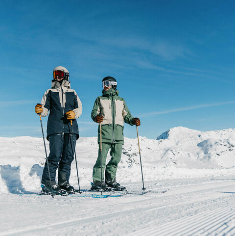 Zwei Skifahrer stehen mit Skiern und Stöcken auf einer präparierten Skipiste vor Berggipfeln.