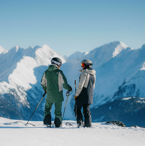 Zwei Skifahrer stehen auf einer Skipiste vor einer verschneiten Berglandschaft.