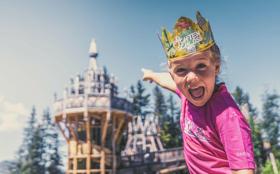 Ein Mädchen trägt eine Papierkrone und zeigt auf das Fichtenschloss.