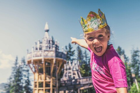 Ein Mädchen trägt eine Papierkrone und zeigt auf das Fichtenschloss.