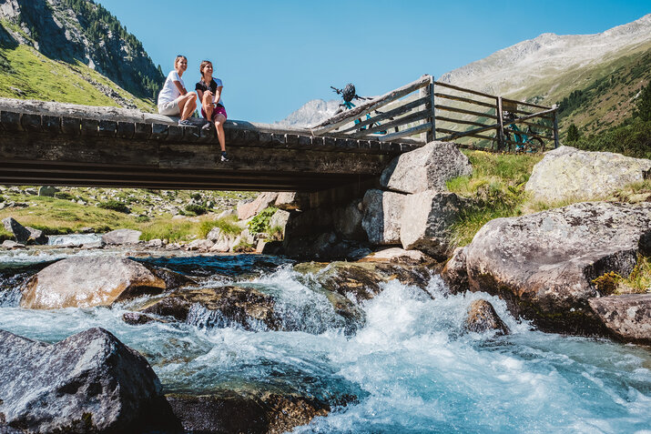 Zwei Frauen sitzen auf einer Brücke auf einem Bach mit Bergkulisse im HIntergrund.