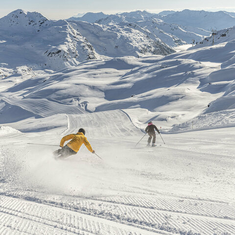 Zwei Skifahrer fahren eine verschneite Bergpiste hinab, umgeben von schneebedeckten Bergen.