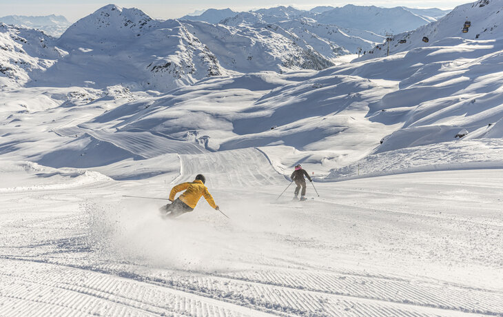 Zwei Skifahrer fahren eine verschneite Bergpiste hinab, umgeben von schneebedeckten Bergen.