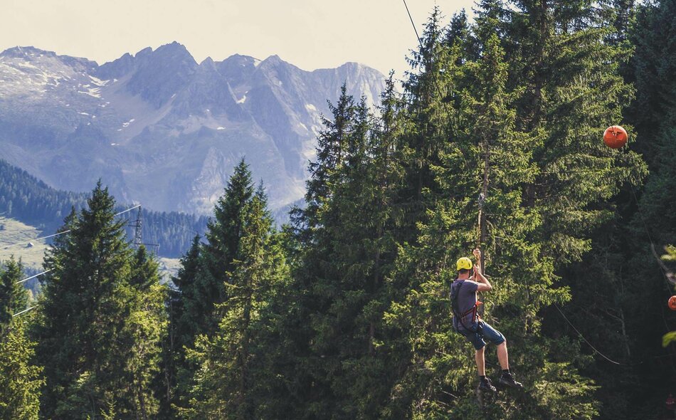 Person fährt mit Helm an einer Flying-Fox-Seilrutsche vor einer Bergkulisse durch den Wald.